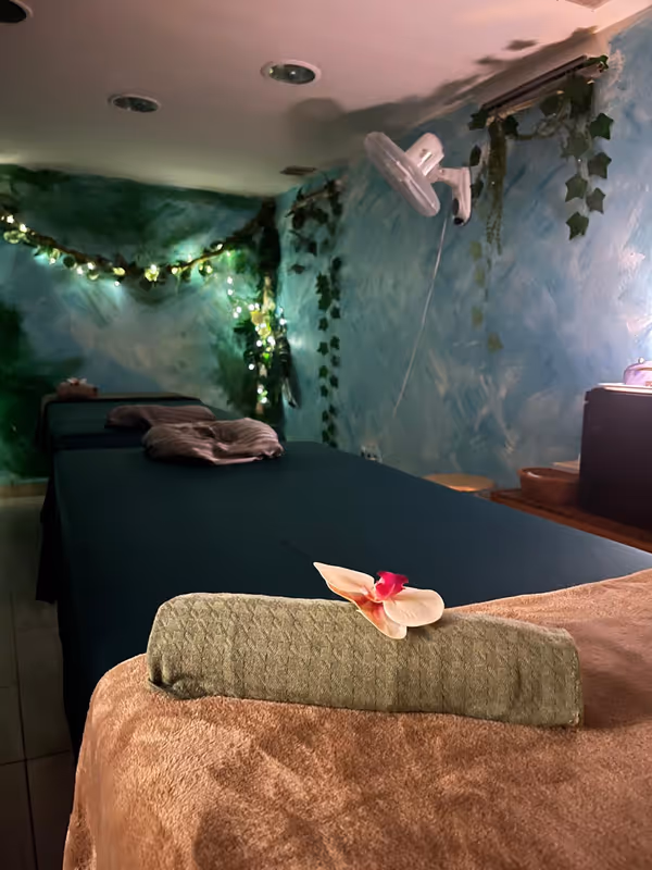 Massage room with green tables and blue walls hung with ivy and fairy lights, and a rolled towel with an orchid